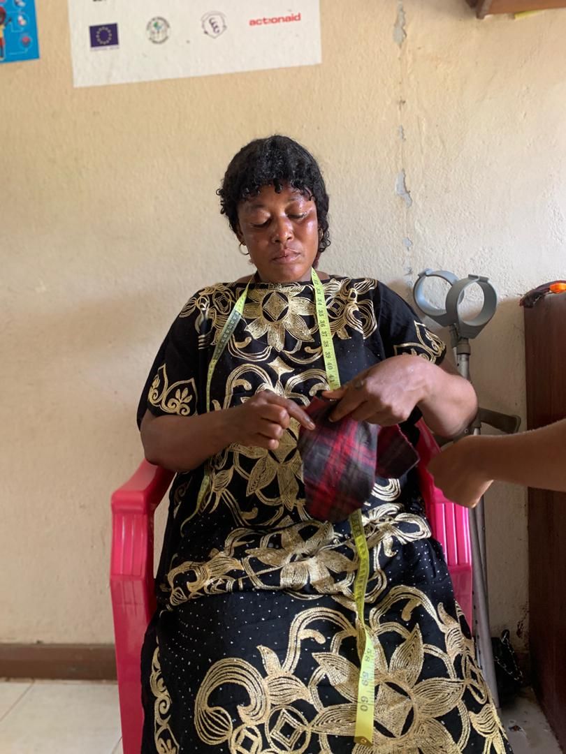A woman with crutches hand-stitching pad fabric with measuring tape around her neck